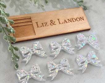 May include: Seven clear, glitter-covered bow-shaped embellishments are arranged near a wooden rectangle engraved with "Liz & Landon." The bows have a shimmering, iridescent effect. Decorative greenery is in the background.