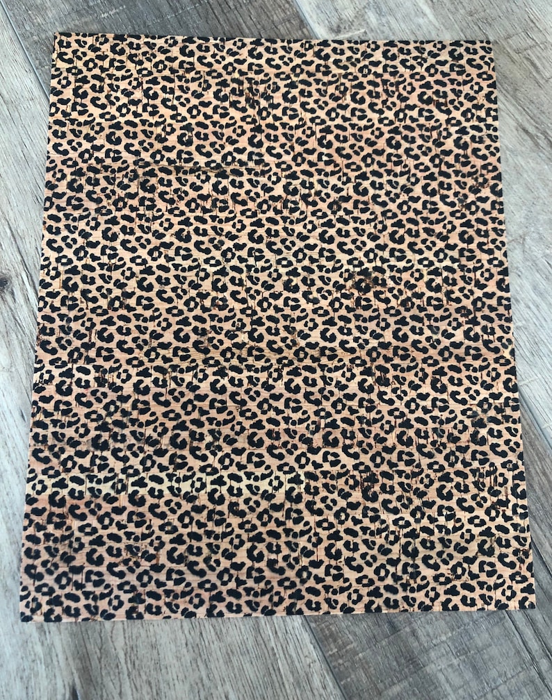 K&ouml;nnte beinhalten: Ein rechteckiges Blatt Kork mit einem Leopardenmuster. Das Muster hat schwarze Flecken auf einem beigefarbenen Hintergrund. Das Korkblatt liegt auf einer grauen Holzoberfl&auml;che.