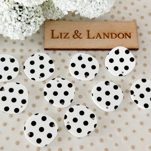May include: Nine white ceramic polka dot buttons with black dots. The buttons have a hole in the top for sewing. The buttons are arranged in a semi-circle on a white background with gold polka dots.