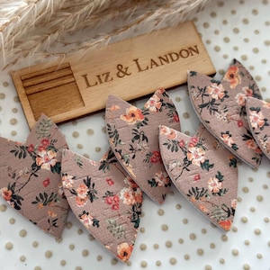 May include: Several heart-shaped cutouts made of a textured, light brown material with a floral pattern featuring orange, pink, and green flowers. A rectangular wooden plaque with the engraved text "LIZ & LANDON" is positioned behind the hearts.