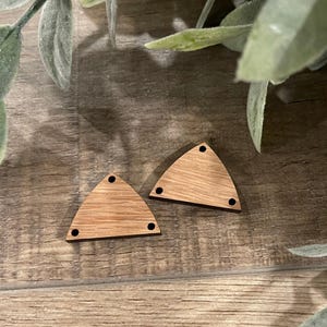 May include: Two unfinished wooden triangle shapes with three small holes and a natural wood grain. The triangles are light brown and sit on a wooden surface with green foliage in the background.