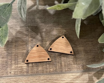 May include: Two unfinished wooden triangle shapes with three small holes and a natural wood grain. The triangles are light brown and sit on a wooden surface with green foliage in the background.