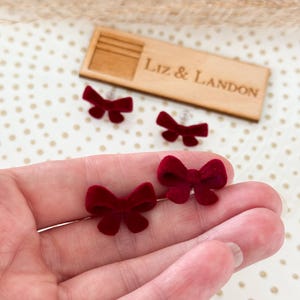 May include: Close-up of two pairs of burgundy velvet bow earrings. The earrings are small and delicate, with a classic bow shape. The earrings are displayed on a hand, with a wooden tag that reads "Liz & Landon" in the background.