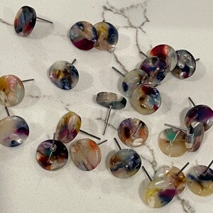 May include: A collection of colorful, round, resin earrings with silver posts. The earrings feature a variety of swirling patterns in shades of blue, purple, white, and gold.