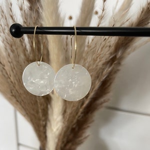 May include: A pair of earrings featuring circular white acrylic pendants with a marbled texture, suspended from thin gold-toned metal hoops. The earrings are displayed hanging from a horizontal black rod, showcasing the translucent quality of the material.