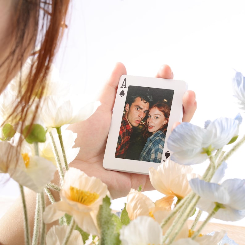 Puede incluir: Una carta de juego blanca, el As de Picas, con una fotograf&iacute;a en color de una pareja. La carta se sostiene frente a un ramo de flores blancas y azules.