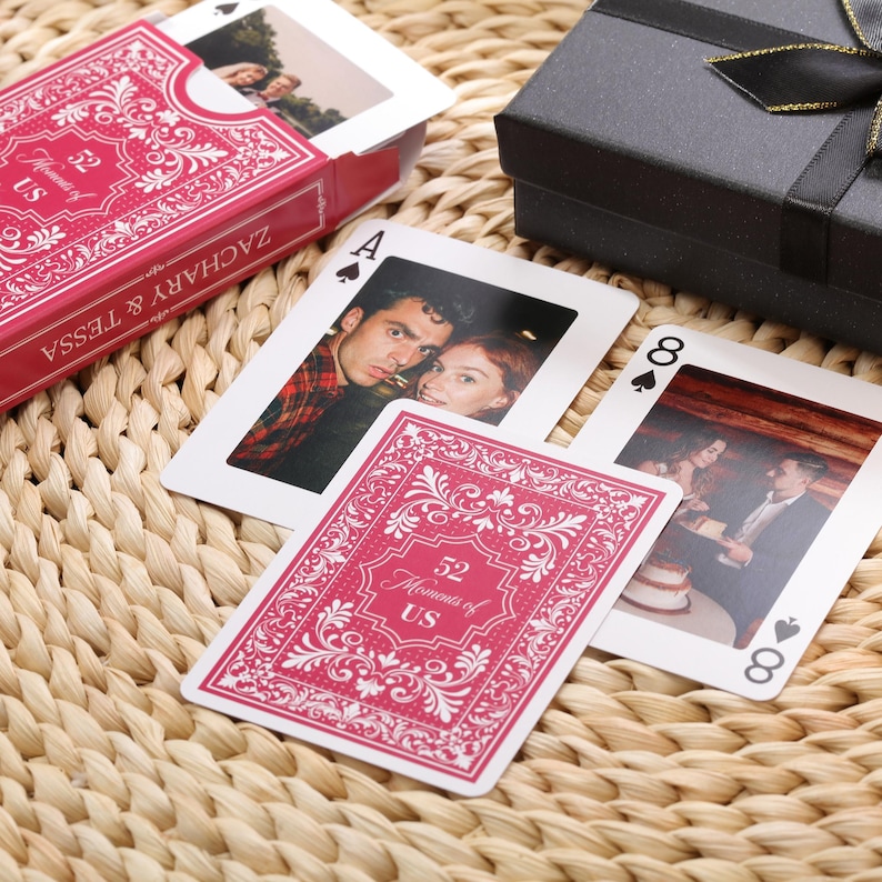 Puede incluir: Una baraja de cartas personalizada con fotos, en una caja roja con el texto "ZACHARY & TESSA" y "52 Moments of US". Las cartas muestran fotos personales y palos de cartas tradicionales.