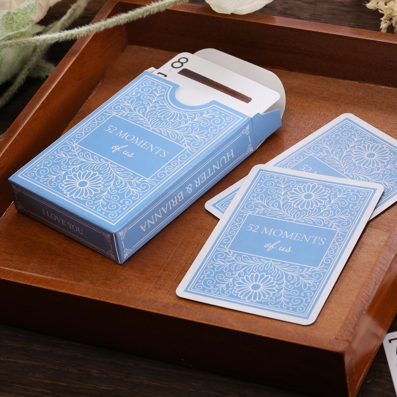 Puede incluir: Una baraja de cartas azul claro con el texto "52 MOMENTS of us" y "HUNTER & BRIANNA" en blanco. La caja y las cartas tienen un dise&ntilde;o floral. Tambi&eacute;n son visibles las palabras "I LOVE YOU".