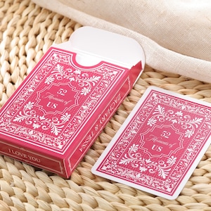 Puede incluir: Una baraja de cartas rosa con adornos blancos y el texto "52 Moments of US". La caja dice "ZACHARY & TESSA" y "I LOVE YOU". Las cartas est&aacute;n sobre una superficie tejida.