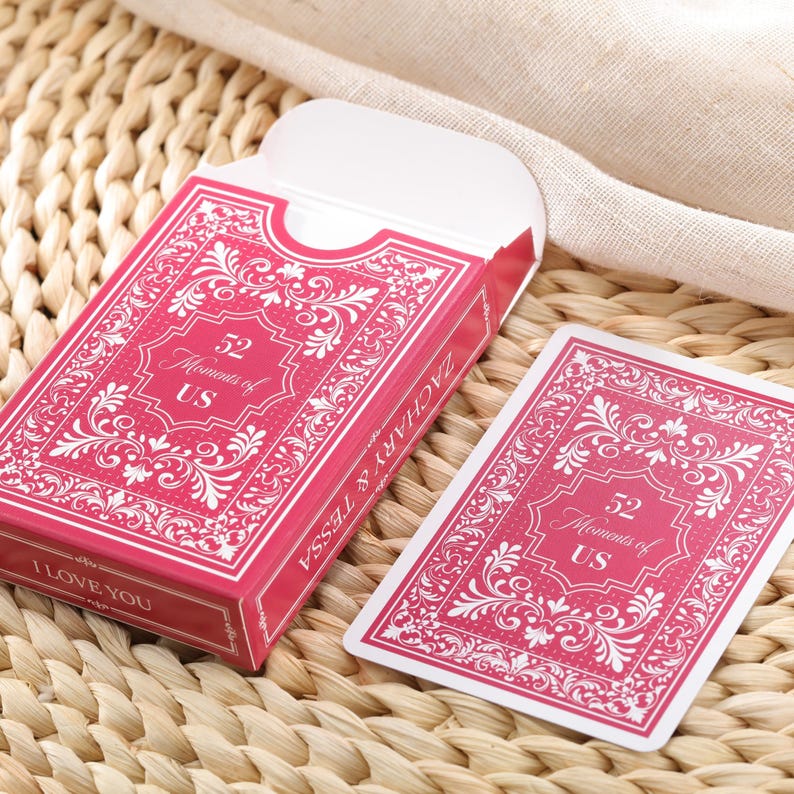 Puede incluir: Una baraja de cartas rosa con adornos blancos y el texto "52 Moments of US". La caja dice "ZACHARY & TESSA" y "I LOVE YOU". Las cartas est&aacute;n sobre una superficie tejida.