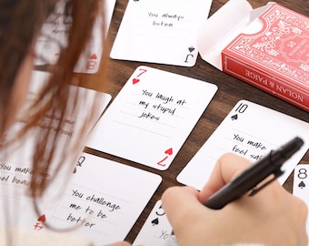 May include: A deck of playing cards with handwritten messages, including "You laugh at my stupid jokes" and "You challenge me to be better." A red and white card box is open, and a hand is writing on a card with a black pen.