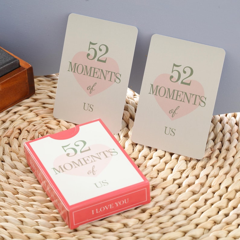 Puede incluir: Una baraja de cartas y dos cartas individuales con el texto "52 Moments of Us" en verde y rosa. La baraja tiene un borde rojo y las palabras "I Love You" en la parte inferior. Las cartas est&aacute;n sobre una superficie tejida.