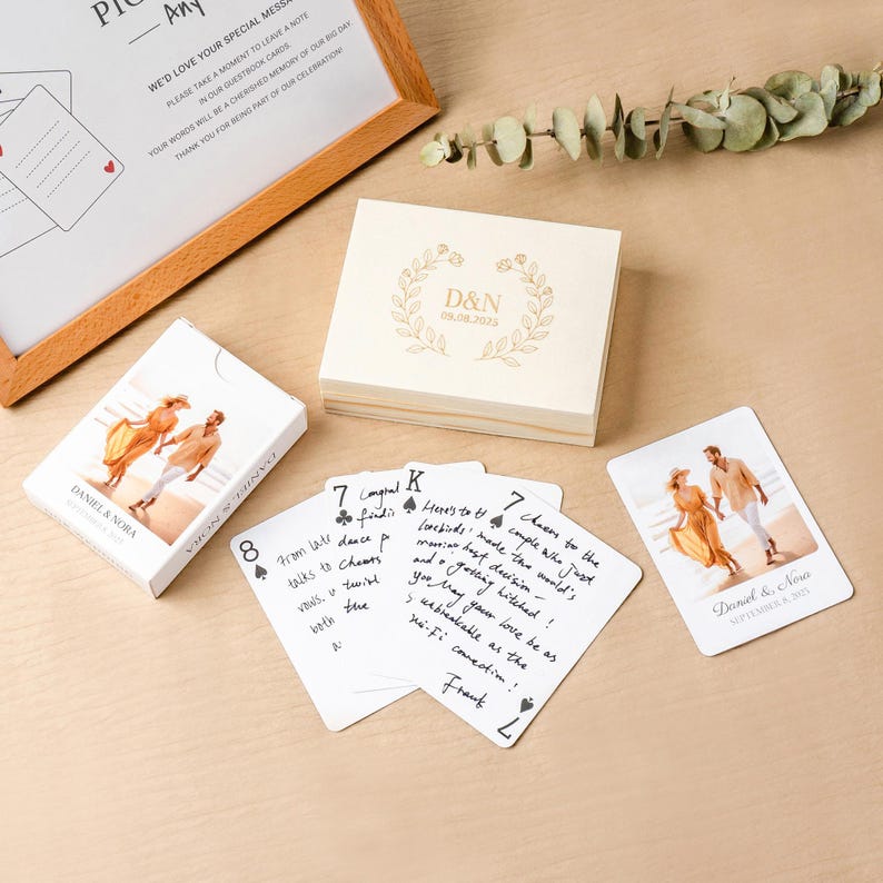 May include: A custom deck of playing cards featuring a photo of a couple on the box and individual cards, a wooden storage box engraved with D&N 08.08.2025, and a framed sign with text inviting guests to leave notes on the cards for the couple.