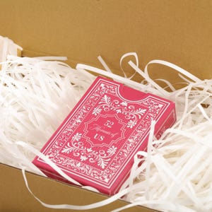 Puede incluir: Una baraja de cartas rosa con dise&ntilde;os florales blancos y el texto "52 Moments of US" est&aacute; metida en relleno de papel blanco dentro de una caja de cart&oacute;n. La baraja tiene un borde adornado.