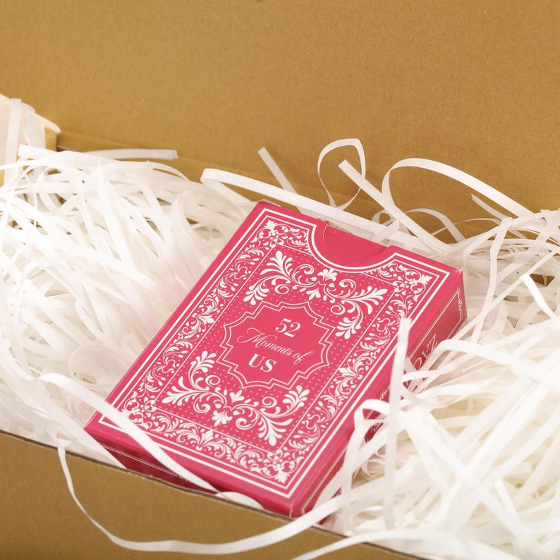 Puede incluir: Una baraja de cartas rosa con dise&ntilde;os florales blancos y el texto "52 Moments of US" est&aacute; metida en relleno de papel blanco dentro de una caja de cart&oacute;n. La baraja tiene un borde adornado.
