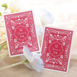 Puede incluir: Dos naipes con fondo rojo y borde blanco adornado. Las cartas presentan el texto "52 Moments of US" en una fuente decorativa. Las cartas se muestran sobre una superficie de color neutro con flores blancas.