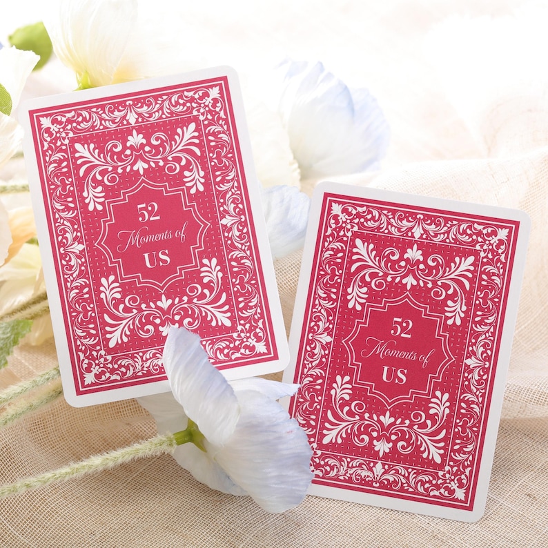 Puede incluir: Dos naipes con fondo rojo y borde blanco adornado. Las cartas presentan el texto "52 Moments of US" en una fuente decorativa. Las cartas se muestran sobre una superficie de color neutro con flores blancas.