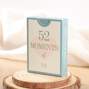 Puede incluir: Una caja de naipes con el texto "52 Moments of Us" en verde y rosa. La caja tiene un borde azul claro y los nombres "Trevor & Kelsey" est&aacute;n impresos en el lateral. La caja se muestra sobre una rodaja de madera.