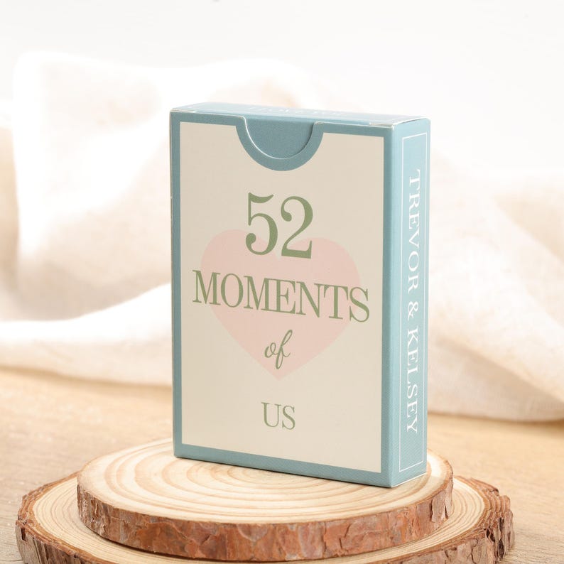 Puede incluir: Una caja de naipes con el texto "52 Moments of Us" en verde y rosa. La caja tiene un borde azul claro y los nombres "Trevor & Kelsey" est&aacute;n impresos en el lateral. La caja se muestra sobre una rodaja de madera.