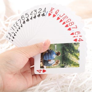 Puede incluir: Un mazo de naipes en abanico, sostenido en una mano. Las cartas presentan palos y n&uacute;meros tradicionales, con una fotograf&iacute;a en la cara del 4 de corazones. La fotograf&iacute;a muestra a dos personas pescando. El fondo es blanco.