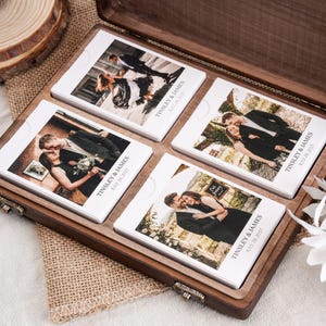 May include: A dark wooden box containing four white rectangular card sets, each featuring a different photograph of a couple and the printed text TINSLEY & JAMES JULY 28, 2027. The cards are arranged in a grid pattern inside the open hinged wooden container.