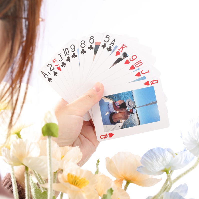 Puede incluir: Una mano sosteniendo una baraja de cartas abanicada. Las cartas muestran varios palos y n&uacute;meros, con una foto personalizada en una carta. El fondo es blanco, con flores en primer plano.