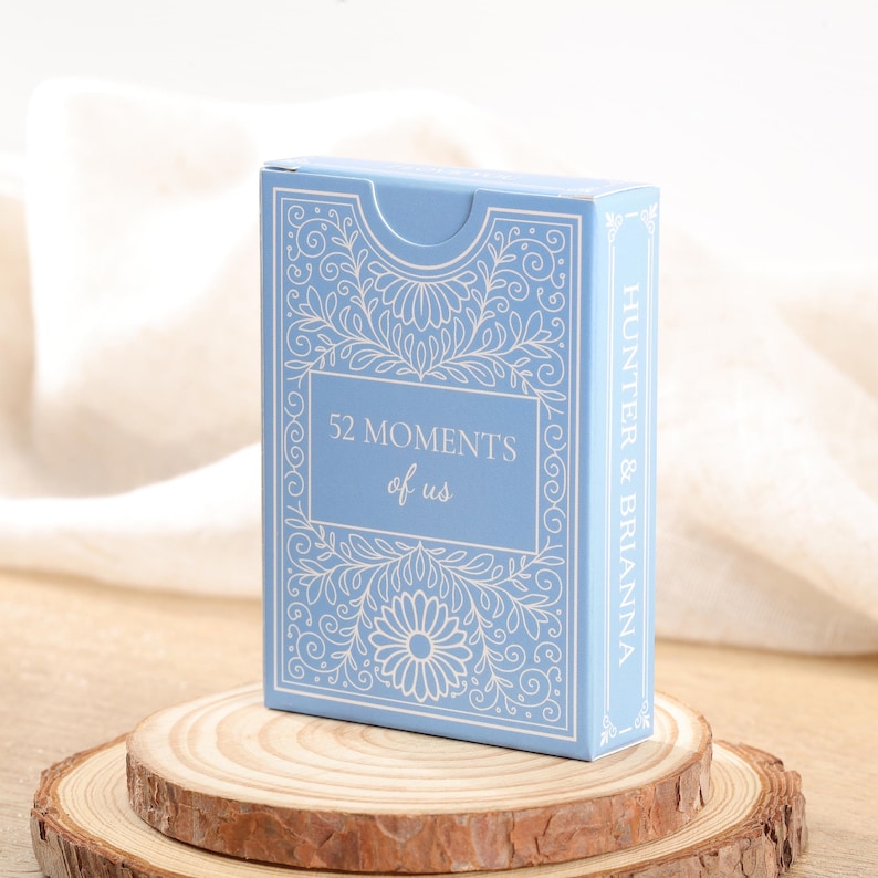 Puede incluir: Una caja de cartas azul claro con dise&ntilde;os florales y de volutas. La caja dice "52 Moments of Us" y "Hunter & Brianna". La caja se muestra sobre una rodaja de madera.