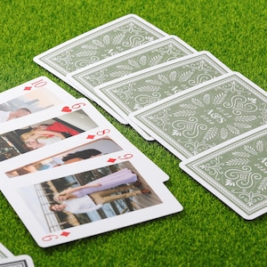 May include: Playing cards with photos on the face and a decorative green and white design on the back. The cards show images of people and the backs have the initials "A&S" and the date "05.25 2026". The cards are arranged on a green surface.