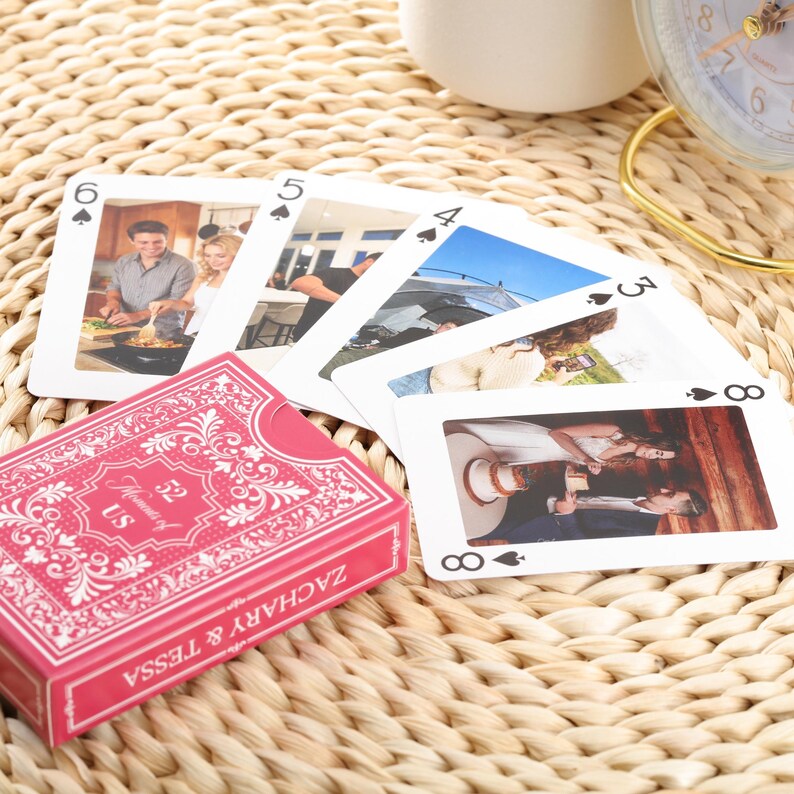 Puede incluir: Una baraja de cartas con una caja rosa con el texto "Moments of US ZACHARY & TESSA". Las cartas muestran fotos y palos de cartas. Las cartas est&aacute;n dispuestas sobre una superficie tejida.