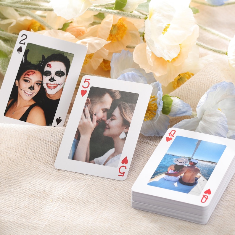 Puede incluir: Una baraja de cartas con fotos personalizadas. Las cartas presentan im&aacute;genes de personas, incluyendo una pareja disfrazada, una pareja abraz&aacute;ndose y una pareja en un barco. Las cartas est&aacute;n dispuestas sobre una superficie beige con flores.