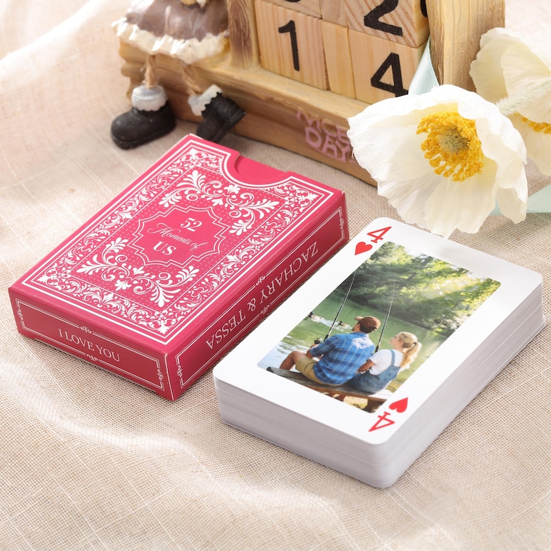 Puede incluir: Una baraja de cartas con una foto personalizada en las caras de las cartas. La caja roja de la baraja dice "52 Moments of US" y "I LOVE YOU". La baraja est&aacute; junto a un calendario de bloques de madera y flores blancas.