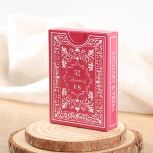 Puede incluir: Una caja de cartas roja con detalles ornamentados blancos y el texto "52 Moments of US". El lateral de la caja dice "ZACHARY & TESSA". La caja se muestra sobre una rodaja de madera.