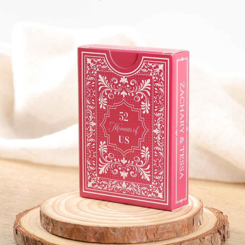 Puede incluir: Una caja de cartas roja con detalles ornamentados blancos y el texto "52 Moments of US". El lateral de la caja dice "ZACHARY & TESSA". La caja se muestra sobre una rodaja de madera.