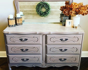 May include: A light pink dresser with six drawers and ornate carvings. The dresser has black drawer pulls and sits on a wooden floor. A mirror with a decorative frame sits above the dresser. A small wreath hangs in the mirror. A vase with dried flowers sits on the dresser.