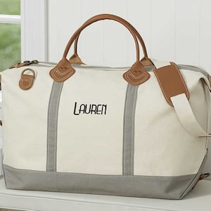 May include: Cream-colored canvas duffel bag with gray accents and brown leather handles and trim. The bag features the name "LAUREN" embroidered in black. Includes a detachable shoulder strap and metal hardware.