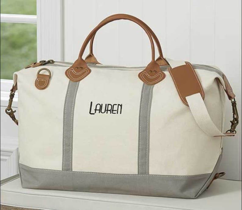 May include: Cream-colored canvas duffel bag with gray accents and brown leather handles and trim. The bag features the name "LAUREN" embroidered in black. Includes a detachable shoulder strap and metal hardware.
