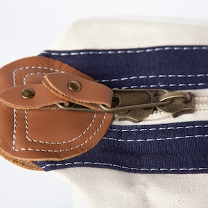 May include: Close-up of a canvas bag with a cream-colored body and navy blue stripes. The bag features a brown leather flap secured by a brass zipper and rivets. White stitching accents the leather.