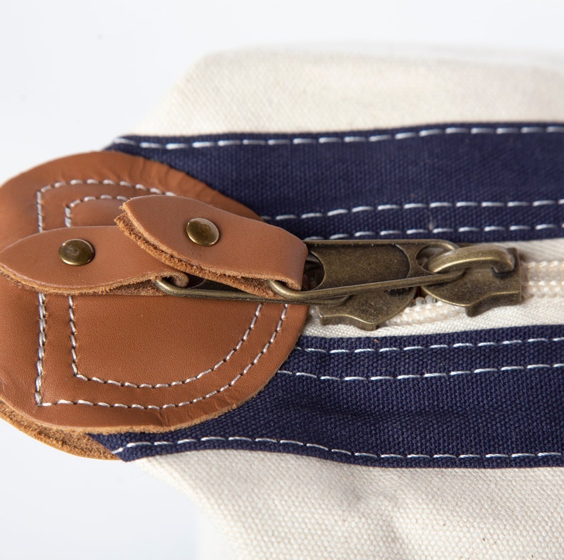 May include: Close-up of a canvas bag with a cream-colored body and navy blue stripes. The bag features a brown leather flap secured by a brass zipper and rivets. White stitching accents the leather.