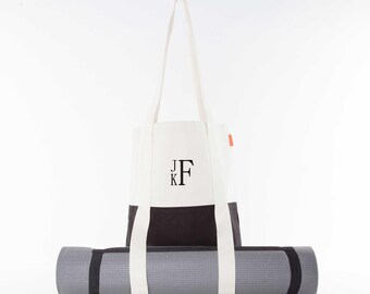 May include: White canvas tote bag with black handles and a black bottom section. The bag has the initials "J.K.F." embroidered on the front. A black and gray yoga mat is in front of the bag.