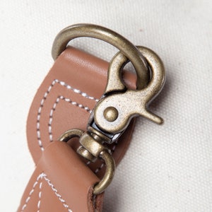 May include: Close-up of a bronze-colored metal clasp attached to a tan leather strap. The leather is stitched with white thread. The background is a light-colored fabric.