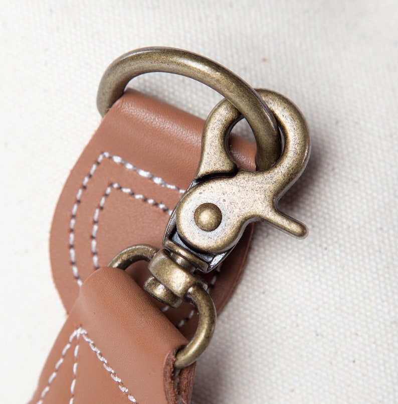 May include: Close-up of a bronze-colored metal clasp attached to a tan leather strap. The leather is stitched with white thread. The background is a light-colored fabric.