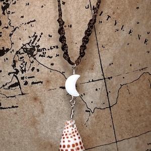 May include: A handcrafted necklace featuring a brown cord with a macrame knot design. The pendant includes a white crescent moon and a seashell with brown spots. The necklace is displayed against a vintage map background.