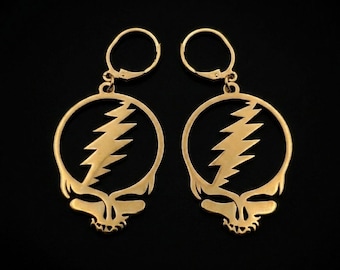 May include: A pair of gold-colored earrings featuring a stylized skull design with a lightning bolt on a black background. The earrings have a circular shape with a hinged closure.