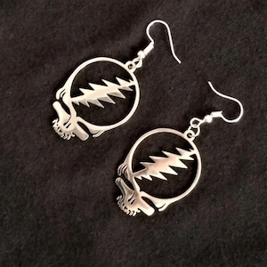 May include: A pair of silver-toned drop earrings featuring a circular frame with a jagged lightning bolt design across the center and a skull-like shape at the base. Each earring is attached to a curved metal hook for pierced ears.