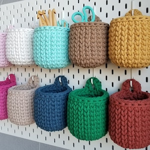 Può includere: Nove cesti appesi all'uncinetto in vari colori, tra cui rosa, bianco, azzurro, marrone, giallo, blu scuro, verde e rosso. I cesti sono appesi a una parete bianca con pannelli forati.