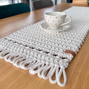 Könnte beinhalten: Ein weißer gehäkelter Tischläufer mit Fransen und einem braunen Lederanhänger. Der Läufer liegt auf einem Holztisch mit einer weißen Tasse und Untertasse mit roten Punkten.