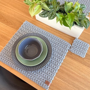 Könnte beinhalten: Ein grauer gehäkelter Tischset mit passendem Untersetzer und einem Satz grauer und grüner Teller und einer Schüssel. Ein weißer rechteckiger Pflanztopf mit grünen Pflanzen befindet sich im Hintergrund. Das Tischset hat einen gewellten Rand.