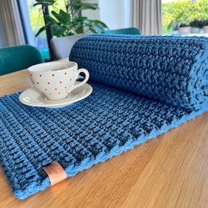 Könnte beinhalten: Ein blauer gehäkelter Tischläufer mit einer weißen Tasse und Untertasse mit roten Punkten darauf. Der Läufer ist auf der rechten Seite des Bildes aufgerollt. Der Läufer hat ein kleines braunes Leder-Etikett mit dem Wort "Blau" darauf.