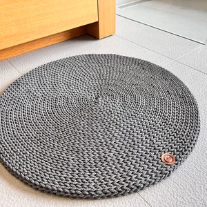 May include: A round, gray, crocheted rug with a brown button in the center. The rug is on a white tiled floor.
