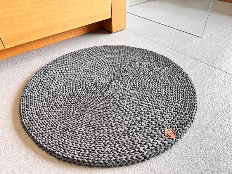 May include: A round, gray, crocheted rug with a brown button in the center. The rug is on a white tiled floor.